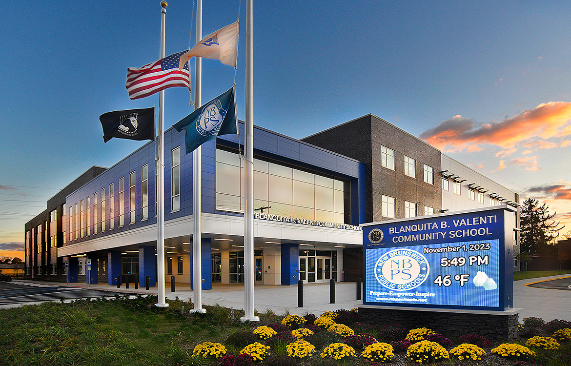 An exterior view of the Blanquita B. Valenti Community School shows the monumnet sign, flags and main entrance. The second floor, elevated on columns, looks into the large glass windows that are a centerpiece of the media center.