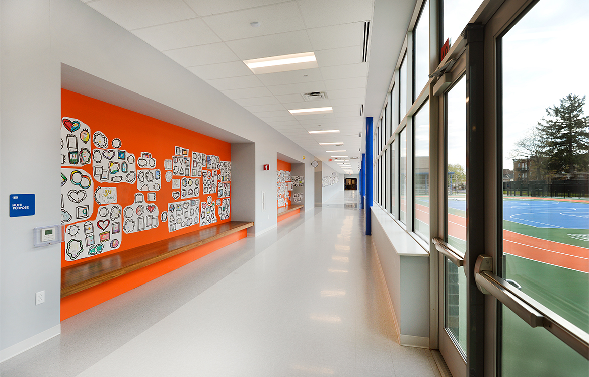 A hallway in the Blanquita B. Valenti Community School overlooks the playground, and also provides benches for socialization and gathering. The walls are painted in bright colors and include student artwork.