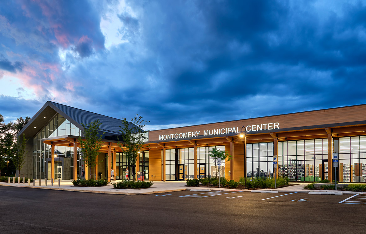The exterior of the Montgomery Municipal Center, photographed at night. Through the windows you can see inside the book stacks and book sorting machine in the library. In the distance, you can council chambers, with a glass facade and a roofline that mimics and gable barn, a nod to the township's agricultural history.