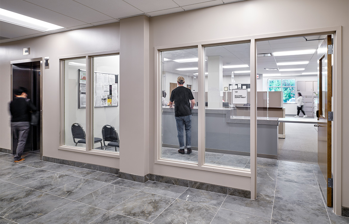 The entrance to the building department through large class windows. Through the windows, a patron is seen being helped at the counter.